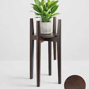 May include: A dark brown wooden plant stand with a round top and four legs. A white ceramic pot with a green plant sits on top. The stand is designed to elevate plants, adding a touch of nature to any space.