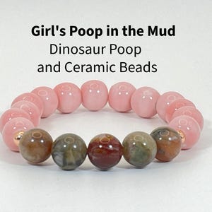 May include: A stretch bracelet featuring a combination of smooth, glossy light pink beads and mottled, earthy brown and green beads. The text Girl's Poop in the Mud Dinosaur Poop and Ceramic Beads is displayed above the bracelet in black font.