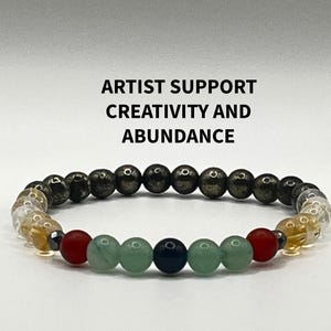 May include: A beaded bracelet with a mix of colors and textures. The bracelet features a variety of beads, including dark gray, clear, red, green, and dark blue. The text "ARTIST SUPPORT CREATIVITY AND ABUNDANCE" is above the bracelet.