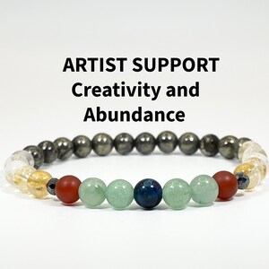 May include: A beaded bracelet with a mix of colors and textures. The bracelet features a variety of beads, including dark gray, clear, yellow, red, green, and blue. Text above the bracelet reads "ARTIST SUPPORT Creativity and Abundance."