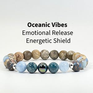 May include: A beaded bracelet featuring a mix of round earth-toned brown, tan, and blue-toned stone beads, accented with silver-colored spacers. The text above the bracelet reads: Oceanic Vibes, Emotional Release, Energetic Shield.