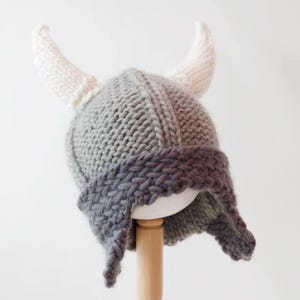 Può includere: Un elmo vichingo lavorato a maglia grigio e bianco con due corna bianche. L'elmo ha paraorecchie ed è esposto su un supporto di legno. Il cappello è realizzato con un filato a maglia grossa.