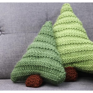 Peut inclure: Deux coussins en forme de sapin de Noël en tricot vert avec des troncs bruns reposent sur un canapé gris rembourré. Les coussins ont un motif texturé et côtelé et sont de tailles différentes. L'arrière-plan est gris neutre.