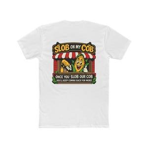 May include: White t-shirt with a colorful graphic featuring a smiling corn character and the text "SLOB ON MY COB" above and "ONCE YOU SLOB OUR COB YOU'LL KEEP COMING BACK FOR MORE!" below. The design is in a cartoon style.