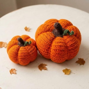 May include: Two crocheted pumpkins in bright orange yarn with dark brown stems and small green curly vines. The pumpkins have a ribbed texture and are arranged with scattered dried brown autumn leaves on a plain white surface.