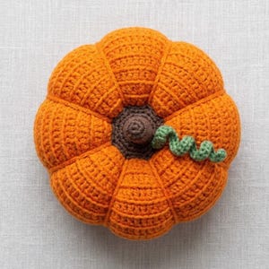 May include: A round, orange crochet pumpkin featuring a dark brown center stem and a single, spiraled green vine attached to the top. The pumpkin is constructed with visible yarn stitches, creating a textured, segmented surface with a soft appearance.