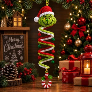 May include: A crocheted hanging ornament featuring a green character head wearing a red and white Santa hat, attached to a red, white, and green spiral body with small dangling spheres, ending in a red and white striped candy cane hook.