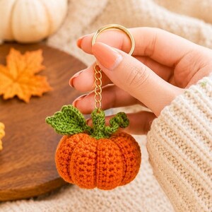 Peut inclure: Un petit porte-clés citrouille orange crocheté, de forme ronde, doté d'une tige verte crochetée et d'une feuille verte. La citrouille est fixée à une chaîne en métal doré et à un anneau fendu circulaire, formant un accessoire fonctionnel.