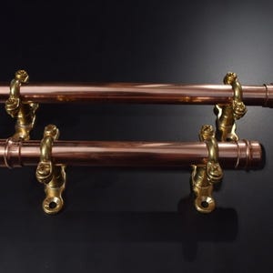 May include: Two copper-coloured towel bars with brass-coloured mounting hardware. The bars are cylindrical and mounted horizontally. The hardware has a decorative design. The background is black.