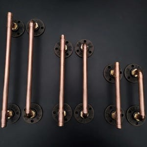 May include: A set of copper pipe door handles in different sizes. Each handle is fixed to a round, black metal base with visible bolt heads. The copper pipes are straight and curved, offering a rustic, industrial style.