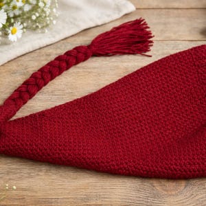 May include: A knitted, deep red gnome hat with a long, braided tail ending in a tassel. The hat is made of a textured fabric and lies on a wooden surface. Daisies and other decorative elements are in the background.