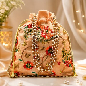 May include: A peach-colored drawstring pouch featuring intricate floral embroidery in shades of red, orange, and green. The bag is adorned with gold-toned beaded trim along the edges and a multi-strand necklace of white pearls draped over the front.