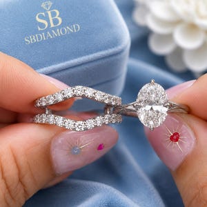 May include: A silver-coloured engagement ring set featuring an oval-cut centre stone and a curved wedding band. Both rings are adorned with small, clear, round-cut stones along the bands. The set is presented in a light blue velvet box with SB SBDIAMOND text.