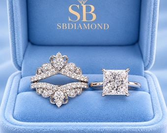 May include: A silver-toned ring set featuring a princess-cut center stone ring and a matching crown-shaped guard ring with pavé-set stones. The jewelry is displayed inside a light blue velvet box with the text SB SBDIAMOND printed in gold.