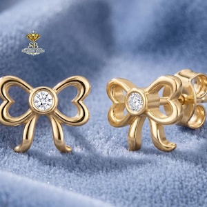 May include: A pair of gold-colored bow-shaped stud earrings. Each earring features a central, round, clear gemstone. The earrings are set against a blue, textured background. The logo "SB Diamond Store" is visible in the upper left corner.