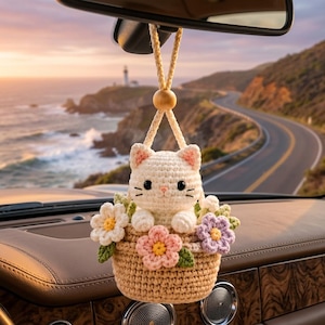 May include: A crochet car hanging ornament featuring a small white cat sitting inside a tan basket. The basket is decorated with three crocheted flowers in white, pink, and purple with green leaves. It hangs from a tan cord with a wooden bead.