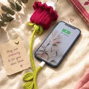 May include: A smartphone connected to a white charging cable encased in a green crocheted stem with leaves, leading to a dark pink crocheted flower cover over the wall plug. A tan note reads: Plug into creativity & let your ideas bloom.