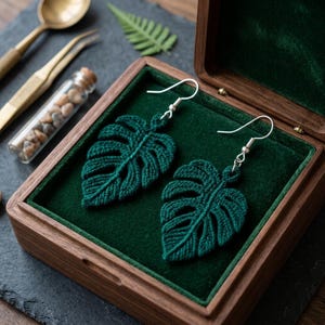 May include: A pair of dark green crochet earrings shaped like monstera leaves with silver-toned metal hooks. The earrings are displayed inside a wooden box lined with dark green velvet, featuring detailed thread work that mimics leaf veins.