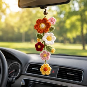 May include: A hanging car accessory featuring a vertical arrangement of crocheted flowers in orange, white, pink, red, and yellow. The flowers are connected by green crocheted stems and leaves, suspended from a small metal bead and a thin cord.