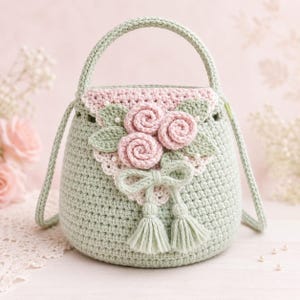 May include: A sage green crochet handbag featuring a triangular flap adorned with three light pink crocheted roses, green leaves, small white beads, and a decorative bow with two matching sage green yarn tassels hanging from the center.