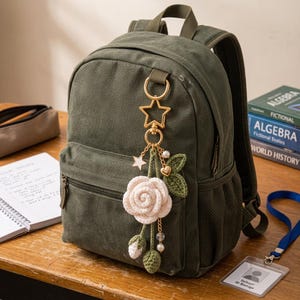May include: An olive green canvas backpack featuring a gold-toned star-shaped keychain. The keychain is adorned with a crocheted white rose, two small green crocheted buds, a gold heart charm, a small star charm, and several white pearl-like beads.