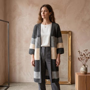 Peut inclure: Un long gilet en crochet ouvert sur le devant, présentant des rayures horizontales en blocs de couleurs gris anthracite foncé, gris clair et crème. Le vêtement possède des manches trois-quarts et un motif texturé à points carrés répétés.