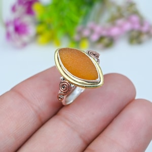 May include: A marquise-shaped orange sea glass ring set in a gold-toned bezel. The band is made of silver-toned metal and features decorative copper and silver-toned wire swirls on the sides, creating a detailed and textured metal finish.
