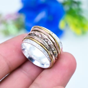 May include: A wide spinner ring featuring multiple stacked bands in silver, gold, and copper tones. The outer silver bands display intricate embossed floral patterns, while the central bands consist of smooth gold and textured copper metal rings.