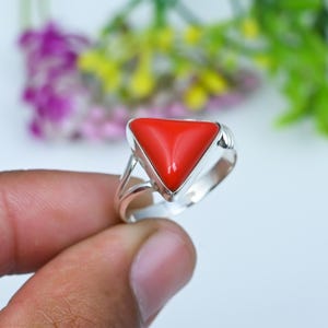 May include: A silver ring featuring a smooth, triangular-shaped red stone set in a polished metal frame. The band has a split design that connects to the base of the triangular setting, creating a clean and simple look for this piece of jewelry.