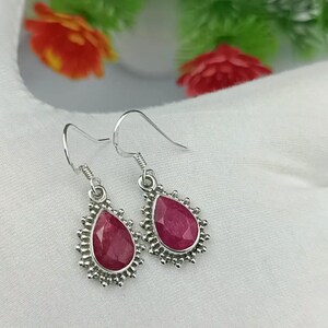 May include: A pair of silver drop earrings featuring faceted, teardrop-shaped dark pink stones. Each stone is set in a decorative silver frame with small bead-like accents along the edge, suspended from simple silver ear wires.