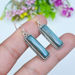 Puede incluir: Un par de pendientes colgantes rectangulares con piedras de labradorita alargadas de iridiscencia azul y gris. Cada piedra está engastada en un marco de metal plateado con borde de cuentas y unida a un gancho de metal plateado simple.