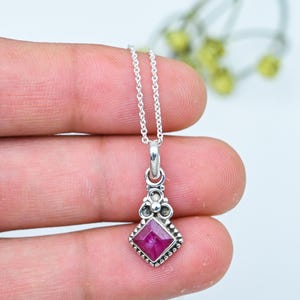 Puede incluir: Un collar con colgante de plata con una piedra preciosa rosa oscuro cuadrada. El colgante tiene un engaste adornado y una cadena delicada. El collar se sostiene en una mano.