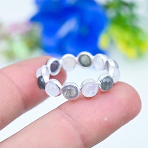 May include: A silver-toned ring featuring a continuous band of alternating round, bezel-set gemstones. The stones display a mix of translucent white, iridescent moonstone-like, and dark grey, speckled cabochons arranged in a circular pattern.