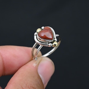 Op de afbeelding: Een zilveren ring met een hartvormige roodbruine steen in een metalen zetting. De zetting bevat twee kleine goudkleurige bolvormige accenten aan de zijkanten van het hart, gemonteerd op een gespleten zilveren band met een gepolijste afwerking.