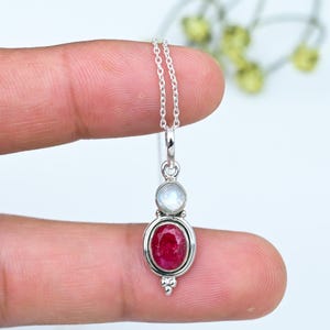 Puede incluir: Un collar con colgante de plata que presenta una piedra preciosa de rubí rojo intenso de forma ovalada y facetada, engastada en un marco de plata pulida, rematada con una piedra lunar redonda e iridiscente, suspendido de una fina cadena.