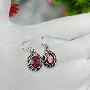 Puede incluir: Un par de pendientes de plata con piedras preciosas facetadas de color rojo rosado intenso y forma ovalada. Cada piedra está engastada en un marco de plata decorativo con cuentas y textura de cuerda trenzada. Ganchos de plata sencillos.