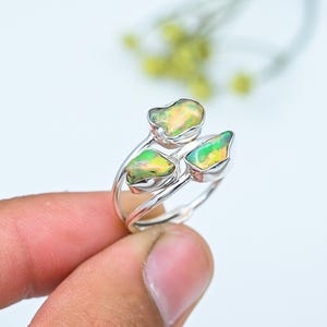 Puede incluir: Un anillo de plata con tres piedras preciosas de ópalo en tonos amarillos y verdes. Las piedras preciosas tienen una forma irregular y están engastadas en una banda de plata pulida. El anillo está sostenido por los dedos de una persona.
