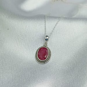 Op de afbeelding: Een zilveren ketting met een ovaalvormige, gefacetteerde donkerroze edelsteen hanger. De steen is gezet in een decoratieve zilveren zetting met een gedraaide touwrand en een kralenrand, hangend aan een fijne zilveren ketting.