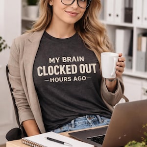 May include: A woman wearing a dark grey t-shirt with the text "MY BRAIN CLOCKED OUT - HOURS AGO-". She is wearing glasses and a beige blazer, holding a white mug. A laptop and notepad are on the desk.