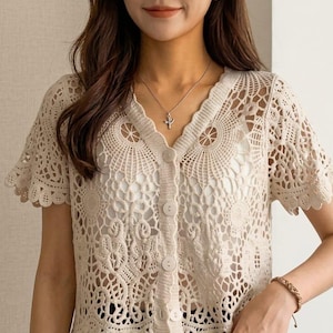 May include: Cream-colored crochet top with a V-neckline and short sleeves. The top features a button-down front and intricate openwork detailing. The garment is made of a soft, lightweight material, perfect for layering or wearing on its own.