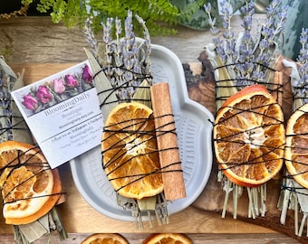 May include: Dried orange slices, cinnamon sticks, and bundles of sage, lavender, and other herbs are arranged on a wooden surface. The bundles are tied with black string and include dried orange slices. A card with the text "BloomingDaily Boutique" is also present.