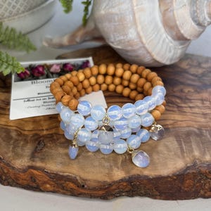 May include: Three bracelets are displayed on a wooden surface. One bracelet is made of light brown wooden beads. The other two bracelets are made of opalescent beads with gold-colored charms and dangling pendants. A small card is visible.