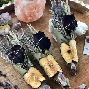 Op de afbeelding: Drie bundels gedroogde kruiden en bloemen gebonden met touw, elk met een gedroogde appelschijf, een zwarte roos en lavendel. De bundels zijn gerangschikt op een houten dienblad met een Himalaya-zoutlamp en amethistkristallen.