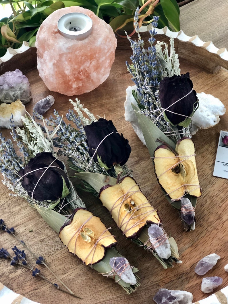 Op de afbeelding: Drie bundels gedroogde kruiden en bloemen gebonden met touw, elk met een gedroogde appelschijf, een zwarte roos en lavendel. De bundels zijn gerangschikt op een houten dienblad met een Himalaya-zoutlamp en amethistkristallen.