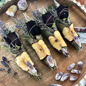 Op de afbeelding: Vijf wierookstokjes met gedroogde appelschijfjes, zwarte rozen, lavendel en salie. Elk wierookstokje is vastgebonden met touw en bevat een amethistkristal.