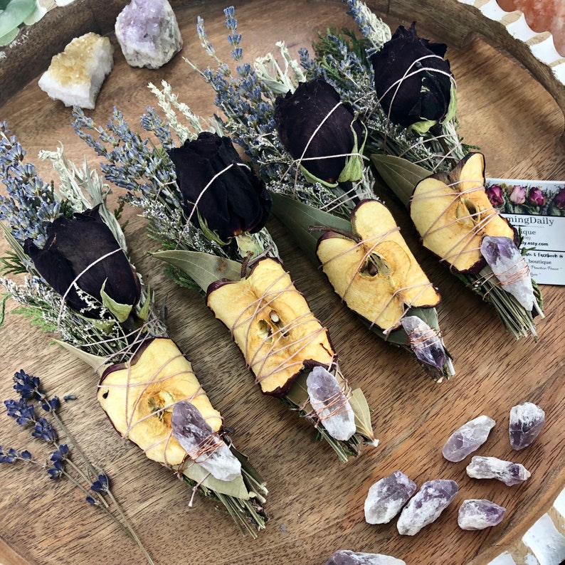 Op de afbeelding: Vijf wierookstokjes met gedroogde appelschijfjes, zwarte rozen, lavendel en salie. Elk wierookstokje is vastgebonden met touw en bevat een amethistkristal.