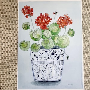 May include: Watercolor painting of a geranium plant in a decorative pot. The plant has bright red flowers and large, round green leaves. The pot is white with a blue floral design. The artwork is signed in the bottom right corner.
