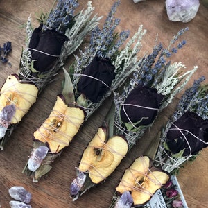 Op de afbeelding: Vijf bundels gedroogde kruiden en bloemen gebonden met touw, waaronder lavendel, salie en gedroogde appelschijfjes. Elke bundel is versierd met een zwarte roos en een klein amethistkristal. De bundels zijn gerangschikt op een houten oppervlak met een klein kaartje waarop "BloominDaily Boutique" staat.