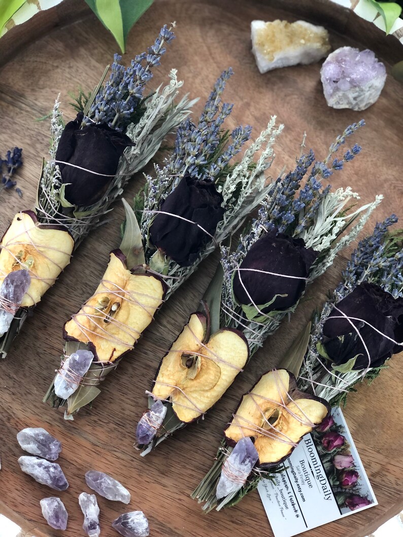 Op de afbeelding: Vijf bundels gedroogde kruiden en bloemen gebonden met touw, waaronder lavendel, salie en gedroogde appelschijfjes. Elke bundel is versierd met een zwarte roos en een klein amethistkristal. De bundels zijn gerangschikt op een houten oppervlak met een klein kaartje waarop "BloominDaily Boutique" staat.