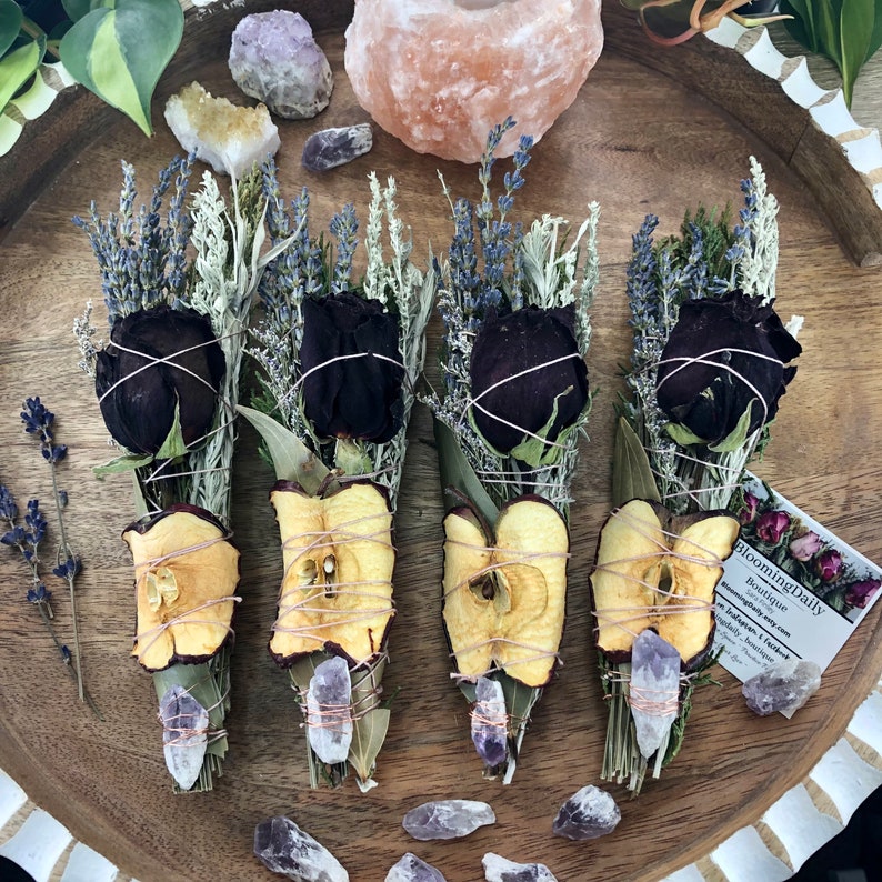 Op de afbeelding: Vier wierookstokjes met gedroogde appelschijfjes, zwarte rozen, lavendel en salie. Elk stokje is vastgebonden met touw en versierd met amethistkristallen. De stokjes zijn gerangschikt op een houten oppervlak met een visitekaartje waarop "Blooming Daily Boutique" staat.